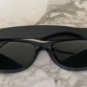 May include: Black Ray-Ban sunglasses with dark lenses. The Ray-Ban logo is visible on the temples. The sunglasses are set against a light-colored, textured background. A classic eyewear accessory.