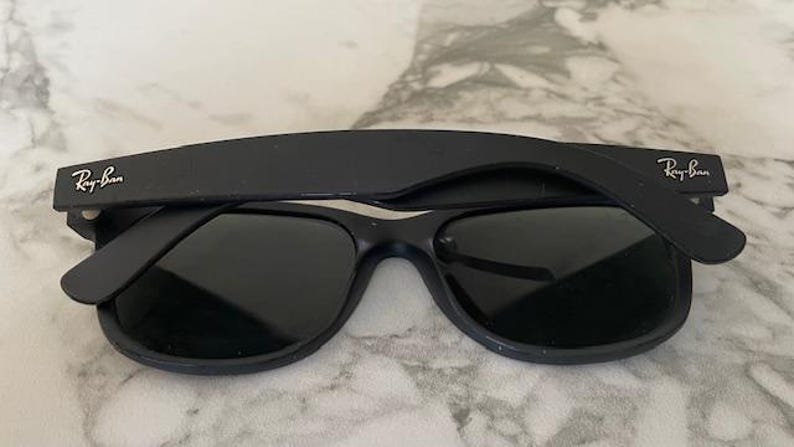 May include: Black Ray-Ban sunglasses with dark lenses. The Ray-Ban logo is visible on the temples. The sunglasses are set against a light-colored, textured background. A classic eyewear accessory.
