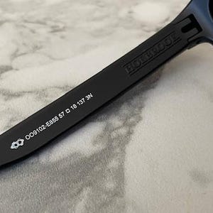 May include: Close-up of black sunglasses with the brand name "HOLBROOK" printed on the arm. The arm also displays the model number OO9102-E855 57 18 137 3N and a small logo. The sunglasses have dark lenses.
