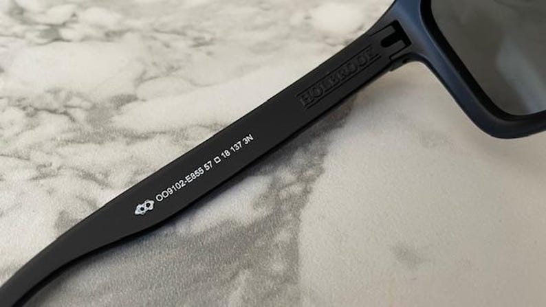 May include: Close-up of black sunglasses with the brand name "HOLBROOK" printed on the arm. The arm also displays the model number OO9102-E855 57 18 137 3N and a small logo. The sunglasses have dark lenses.