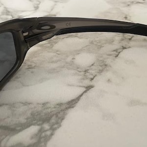 May include: Close-up of a pair of dark gray sunglasses. The sunglasses have a curved frame and dark lenses. The arms of the sunglasses are black and extend to the ear. The sunglasses are resting on a marble surface.