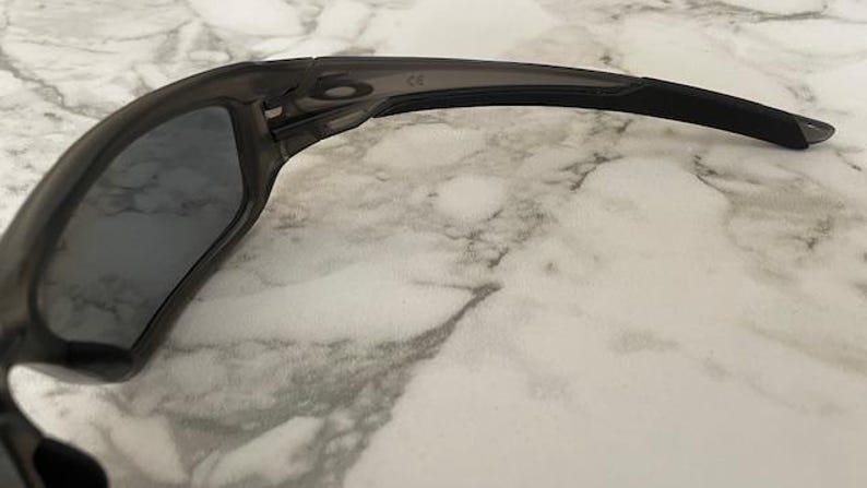 May include: Close-up of a pair of dark gray sunglasses. The sunglasses have a curved frame and dark lenses. The arms of the sunglasses are black and extend to the ear. The sunglasses are resting on a marble surface.