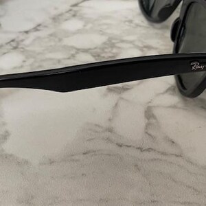 K&ouml;nnte beinhalten: Schwarze Sonnenbrille mit dunklen Gl&auml;sern. Der Rahmen ist gl&auml;nzend schwarz mit dem Ray-Ban-Logo auf dem B&uuml;gel. Die Sonnenbrille liegt auf einer wei&szlig;-grauen Marmoroberfl&auml;che.
