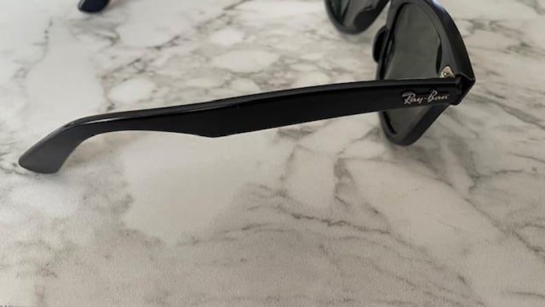 K&ouml;nnte beinhalten: Schwarze Sonnenbrille mit dunklen Gl&auml;sern. Der Rahmen ist gl&auml;nzend schwarz mit dem Ray-Ban-Logo auf dem B&uuml;gel. Die Sonnenbrille liegt auf einer wei&szlig;-grauen Marmoroberfl&auml;che.