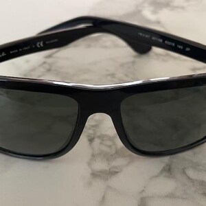 Puede incluir: Gafas de sol negras con lentes oscuras. Las gafas tienen una montura rectangular cl&aacute;sica con un acabado brillante. El logotipo de Ray-Ban es visible en la lente izquierda. Las gafas descansan sobre una superficie de m&aacute;rmol.