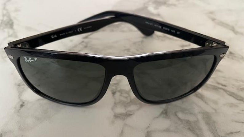 Puede incluir: Gafas de sol negras con lentes oscuras. Las gafas tienen una montura rectangular cl&aacute;sica con un acabado brillante. El logotipo de Ray-Ban es visible en la lente izquierda. Las gafas descansan sobre una superficie de m&aacute;rmol.
