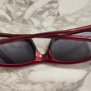 May include: Red-framed sunglasses with dark lenses. The sunglasses have a glossy finish and a curved design. The arms of the sunglasses are folded over the lenses. The lenses are a dark gray color.