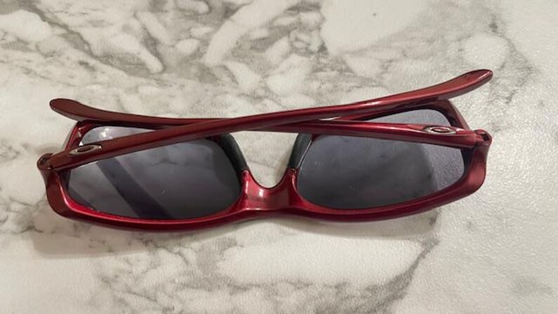 May include: Red-framed sunglasses with dark lenses. The sunglasses have a glossy finish and a curved design. The arms of the sunglasses are folded over the lenses. The lenses are a dark gray color.
