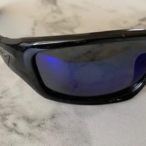 May include: Black sunglasses with a curved frame and blue-tinted lenses. The sunglasses have a sleek, modern design, suitable for outdoor activities. The lenses reflect a blue hue.