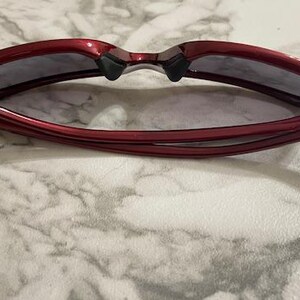 May include: Red sunglasses with dark lenses are displayed open, resting on a marble surface. The frames are a glossy, deep red, and the lenses are a dark gray. The arms of the sunglasses are curved and extend outward.