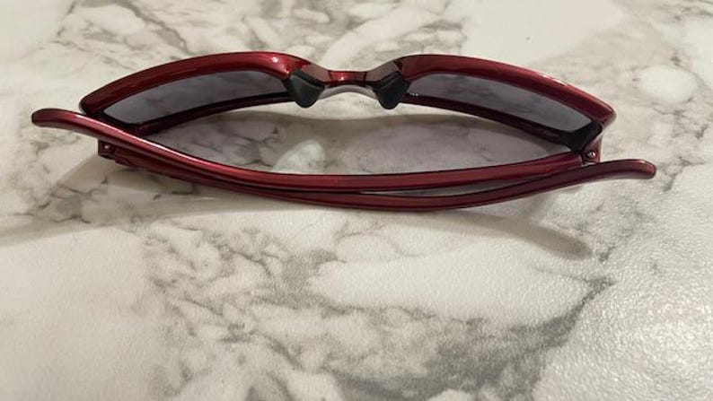 May include: Red sunglasses with dark lenses are displayed open, resting on a marble surface. The frames are a glossy, deep red, and the lenses are a dark gray. The arms of the sunglasses are curved and extend outward.