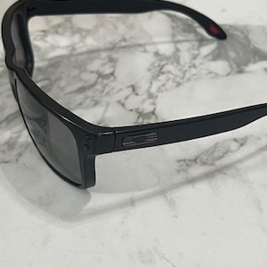 May include: Black sunglasses with a rectangular frame and dark lenses. The temples have a small red accent. The sunglasses are set against a white and gray marble background.