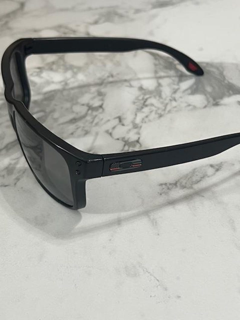 May include: Black sunglasses with a rectangular frame and dark lenses. The temples have a small red accent. The sunglasses are set against a white and gray marble background.