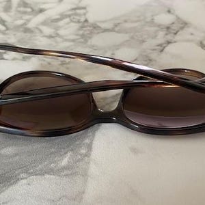 May include: Brown tortoiseshell sunglasses with dark lenses. The sunglasses have a classic design with a rounded frame and a sleek, modern look. The arms are folded, resting on a light-colored surface.