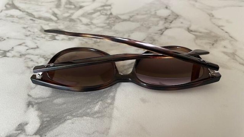 May include: Brown tortoiseshell sunglasses with dark lenses. The sunglasses have a classic design with a rounded frame and a sleek, modern look. The arms are folded, resting on a light-colored surface.