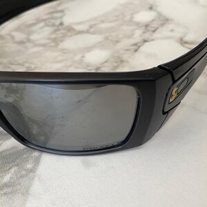 May include: Black wraparound sunglasses with grey reflective lenses. The thick frames feature a small gold emblem on the temple. The sunglasses are designed for optimal eye protection.