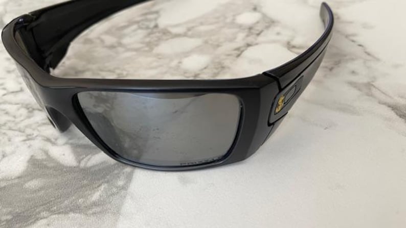 May include: Black wraparound sunglasses with grey reflective lenses. The thick frames feature a small gold emblem on the temple. The sunglasses are designed for optimal eye protection.