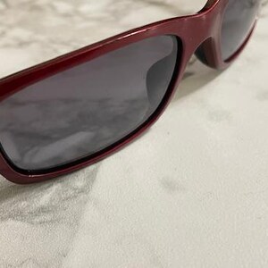 May include: Close-up of burgundy sunglasses with dark lenses. The sunglasses have a rectangular frame and are angled slightly to the right. The background is a light-colored, textured surface.