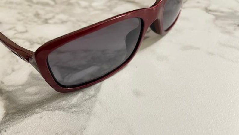 May include: Close-up of burgundy sunglasses with dark lenses. The sunglasses have a rectangular frame and are angled slightly to the right. The background is a light-colored, textured surface.