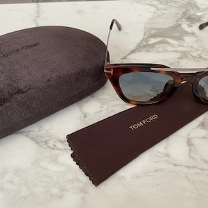 以下が含まれることがあります： 青みがかったレンズのべっ甲柄サングラス、茶色のケース、クリーニングクロス。 サングラスは特徴的なキャットアイシェイプで、クロスにはブランド名「TOM FORD」が見えます。
