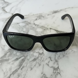May include: Black sunglasses with a curved frame and dark lenses. The sunglasses have a subtle cat-eye shape and the Ray-Ban logo is visible on the left lens. The glasses are set against a marble background.