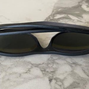 May include: Black sunglasses with dark lenses, folded in half. The sunglasses have a matte finish and a classic design. The Ray-Ban logo is visible on the temple. The sunglasses are resting on a light-colored surface.