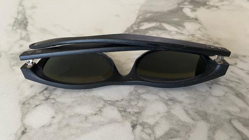 May include: Black sunglasses with dark lenses, folded in half. The sunglasses have a matte finish and a classic design. The Ray-Ban logo is visible on the temple. The sunglasses are resting on a light-colored surface.