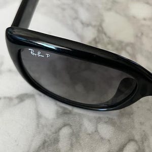K&ouml;nnte beinhalten: Schwarze Sonnenbrille mit dunkelgrauen Gl&auml;sern. Das Ray-Ban-Logo ist auf dem linken Glas zu sehen. Die Sonnenbrille hat ein schlankes, modernes Design mit gl&auml;nzendem Finish.
