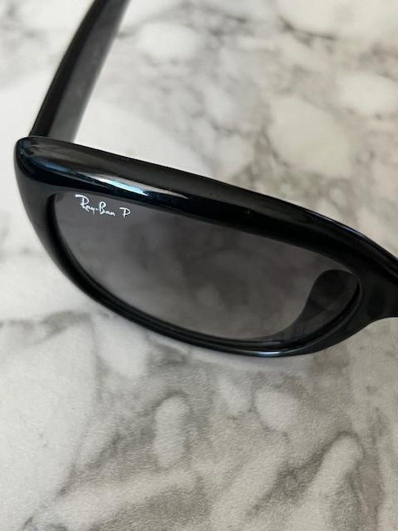 K&ouml;nnte beinhalten: Schwarze Sonnenbrille mit dunkelgrauen Gl&auml;sern. Das Ray-Ban-Logo ist auf dem linken Glas zu sehen. Die Sonnenbrille hat ein schlankes, modernes Design mit gl&auml;nzendem Finish.