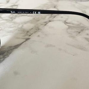 Puede incluir: Gafas de sol negras con lentes oscuras. La montura es negra y tiene el texto "MADE IN ITALY" impreso en la patilla. Las gafas de sol est&aacute;n sobre una superficie de m&aacute;rmol blanco y gris.