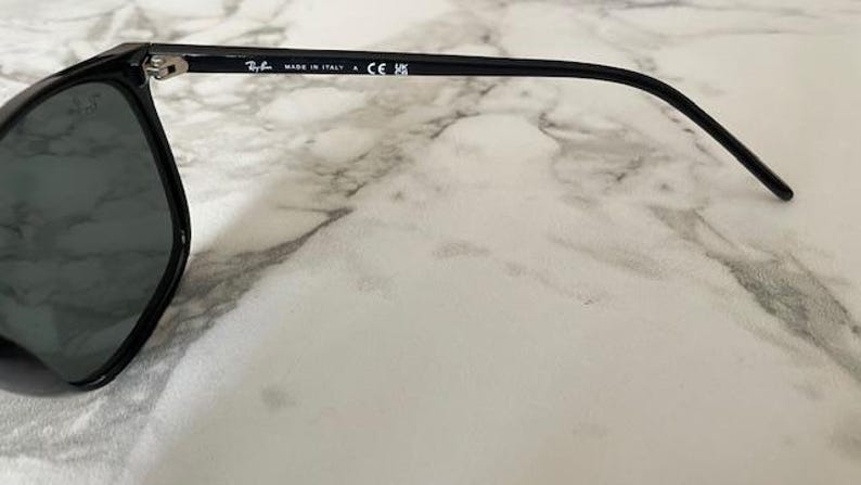 Puede incluir: Gafas de sol negras con lentes oscuras. La montura es negra y tiene el texto "MADE IN ITALY" impreso en la patilla. Las gafas de sol est&aacute;n sobre una superficie de m&aacute;rmol blanco y gris.
