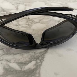 May include: Black sunglasses with a glossy finish. The sunglasses have a curved frame and dark lenses. The arms of the sunglasses are folded. The sunglasses are resting on a white and gray marble surface.