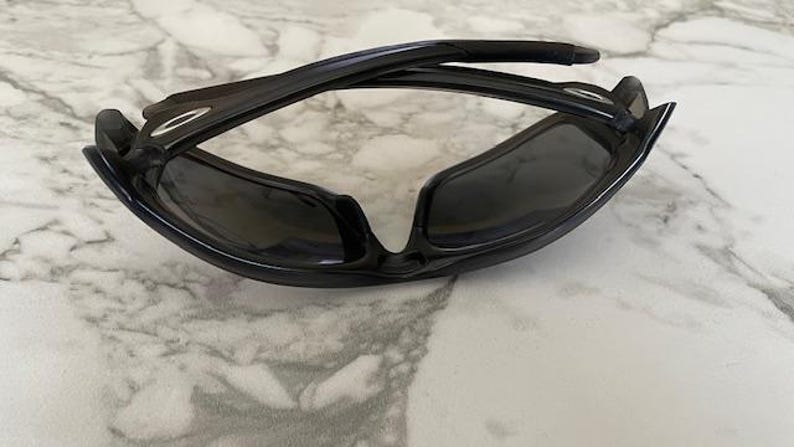 May include: Black sunglasses with a glossy finish. The sunglasses have a curved frame and dark lenses. The arms of the sunglasses are folded. The sunglasses are resting on a white and gray marble surface.