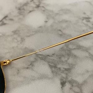 May include: Close-up of sunglasses with a gold-colored metal frame and dark lenses. The temple arm is thin and gold, transitioning to a black tip. The lenses have a dark tint, and the frame is detailed with small text.