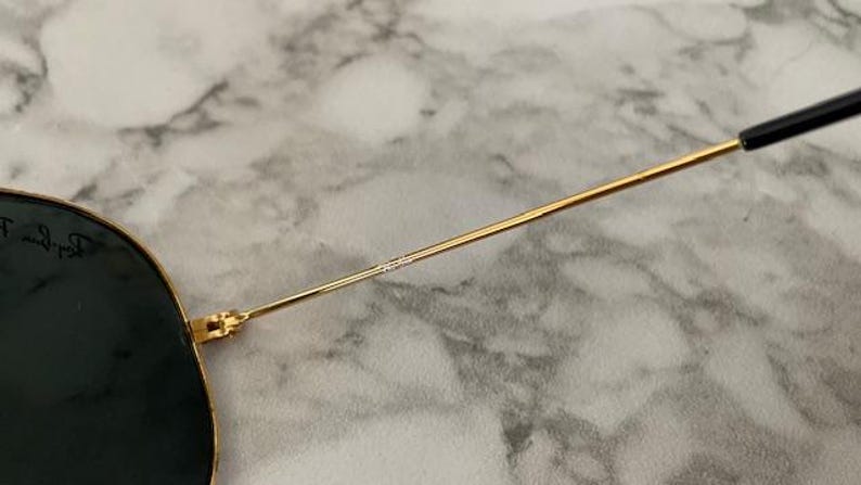May include: Close-up of sunglasses with a gold-colored metal frame and dark lenses. The temple arm is thin and gold, transitioning to a black tip. The lenses have a dark tint, and the frame is detailed with small text.