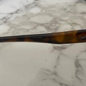 Op de afbeelding: Close-up van een zonnebril met een schildpadpatroon op de pootjes. De pootjes zijn donkerbruin met een lichter bruin en geel patroon. De zonnebril heeft een strak, modern design.