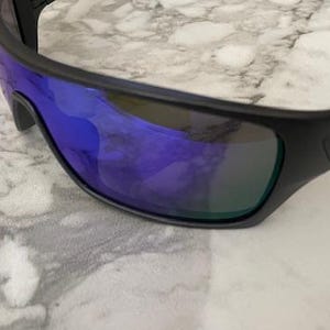 Może przedstawiać: Czarne okulary przeciwsłoneczne z niebieskimi lustrzanymi soczewkami. Okulary mają elegancki, sportowy design z matową czarną oprawką. Soczewki odbijają żywy niebiesko-zielony odcień, oferując stylowy i funkcjonalny dodatek.