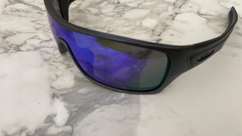 Może przedstawiać: Czarne okulary przeciwsłoneczne z niebieskimi lustrzanymi soczewkami. Okulary mają elegancki, sportowy design z matową czarną oprawką. Soczewki odbijają żywy niebiesko-zielony odcień, oferując stylowy i funkcjonalny dodatek.