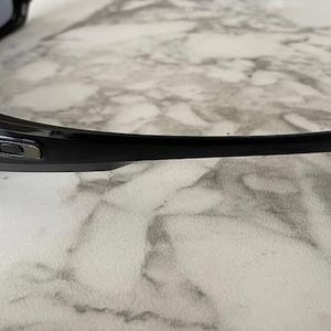 K&ouml;nnte beinhalten: Schwarze Sonnenbrille mit gebogenem B&uuml;gel. Die Sonnenbrille hat eine gl&auml;nzende Oberfl&auml;che und ein silbernes Logo auf dem Rahmen. Die Gl&auml;ser sind dunkel, und die Sonnenbrille liegt auf einer Marmoroberfl&auml;che.