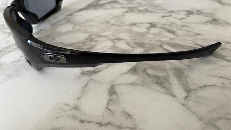 K&ouml;nnte beinhalten: Schwarze Sonnenbrille mit gebogenem B&uuml;gel. Die Sonnenbrille hat eine gl&auml;nzende Oberfl&auml;che und ein silbernes Logo auf dem Rahmen. Die Gl&auml;ser sind dunkel, und die Sonnenbrille liegt auf einer Marmoroberfl&auml;che.