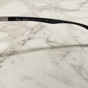 May include: Black Ray-Ban sunglasses with blue-tinted lenses. The frame is black with the text "LITEFORCE MADE IN ITALY P CE POLARIZED RB 4305" printed on the temple. The sunglasses are on a white marble surface.