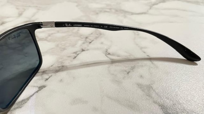 May include: Black Ray-Ban sunglasses with blue-tinted lenses. The frame is black with the text "LITEFORCE MADE IN ITALY P CE POLARIZED RB 4305" printed on the temple. The sunglasses are on a white marble surface.