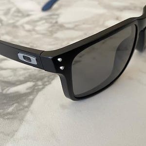 May include: Black sunglasses with gray lenses. The frames are black with a silver logo on the temples. The arms have blue accents. The sunglasses are resting on a white and gray marble surface.