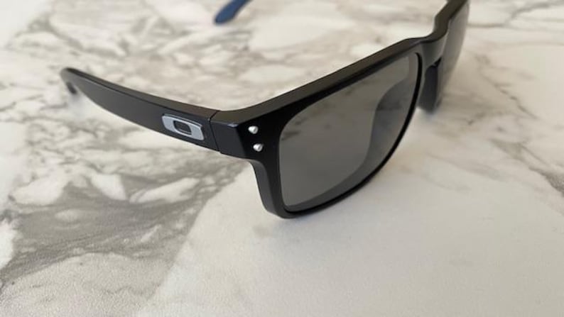 May include: Black sunglasses with gray lenses. The frames are black with a silver logo on the temples. The arms have blue accents. The sunglasses are resting on a white and gray marble surface.