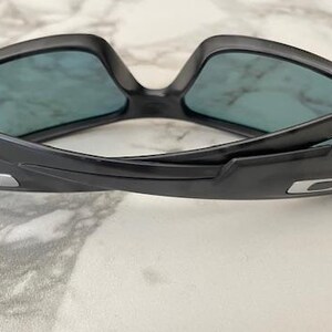 Puede incluir: Gafas de sol negras con lentes azules. Las gafas tienen un dise&ntilde;o moderno con una montura curvada y un logotipo plateado en las patillas. Las lentes son rectangulares y ofrecen una visi&oacute;n clara.