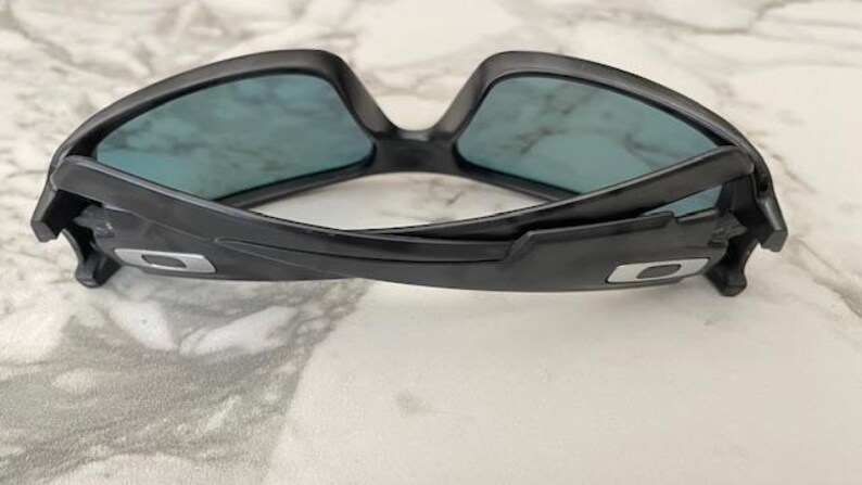 Puede incluir: Gafas de sol negras con lentes azules. Las gafas tienen un dise&ntilde;o moderno con una montura curvada y un logotipo plateado en las patillas. Las lentes son rectangulares y ofrecen una visi&oacute;n clara.