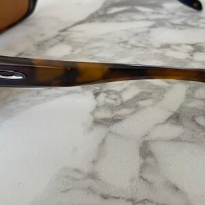 Op de afbeelding: Close-up van een zonnebril met een donkerbruin montuur en glazen. De pootjes hebben een schildpadpatroon. Het Oakley-logo is zichtbaar op de poot. De zonnebril ligt op een wit en grijs marmeren oppervlak.