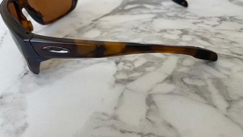 Op de afbeelding: Close-up van een zonnebril met een donkerbruin montuur en glazen. De pootjes hebben een schildpadpatroon. Het Oakley-logo is zichtbaar op de poot. De zonnebril ligt op een wit en grijs marmeren oppervlak.