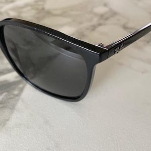 Puede incluir: Gafas de sol negras con lentes oscuras. Las monturas son rectangulares y brillantes, con un peque&ntilde;o logotipo en la patilla. Las gafas de sol est&aacute;n sobre un fondo claro.