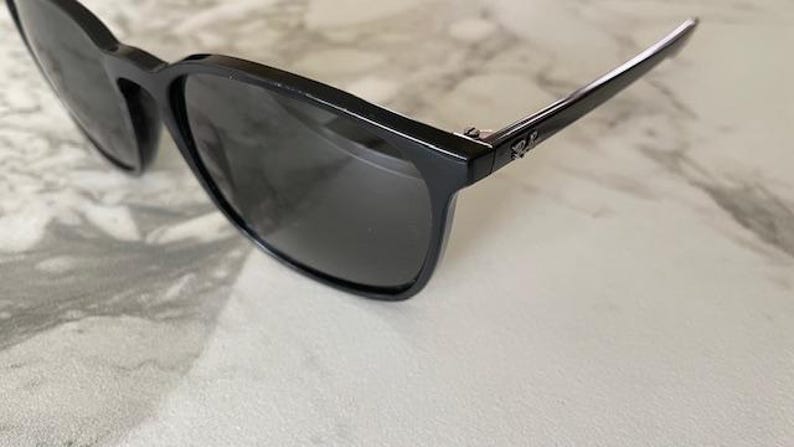 Puede incluir: Gafas de sol negras con lentes oscuras. Las monturas son rectangulares y brillantes, con un peque&ntilde;o logotipo en la patilla. Las gafas de sol est&aacute;n sobre un fondo claro.